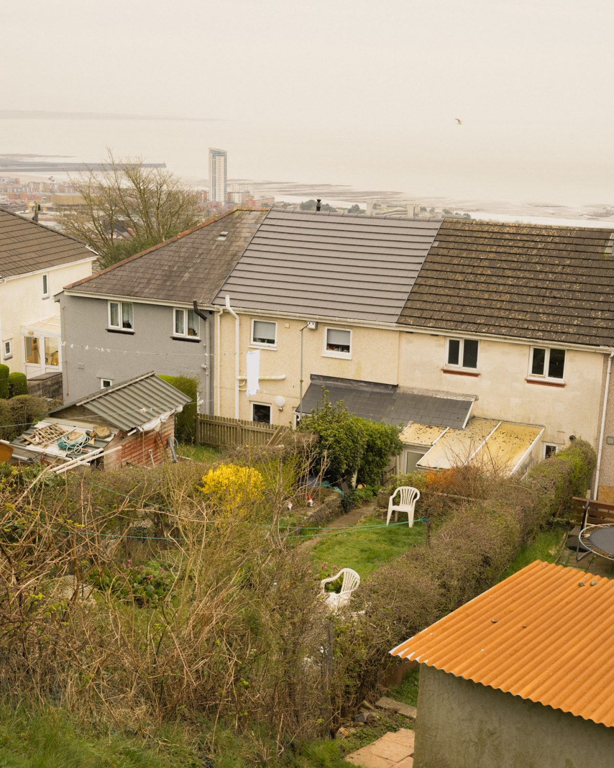 View of a suburban backyard with green bushes, outdoor chairs, sheds, and houses; the sea is visible in the background under a cloudy sky.