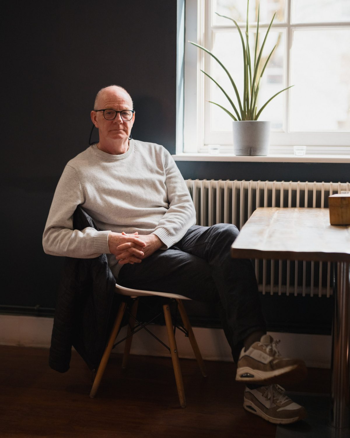 A man with glasses and a grey jumper sat at a table posing for the camera in front of a potted plant