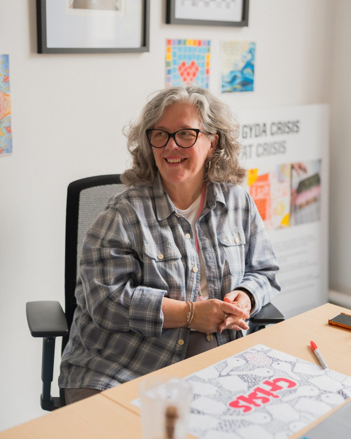 A woman with glasses sat at a desk with pens and a hand drawn poster
