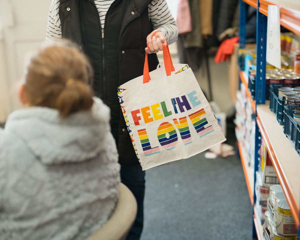 A woman holding a white bag with the words "Feel the Love" written on it. The bag has a rainbow design